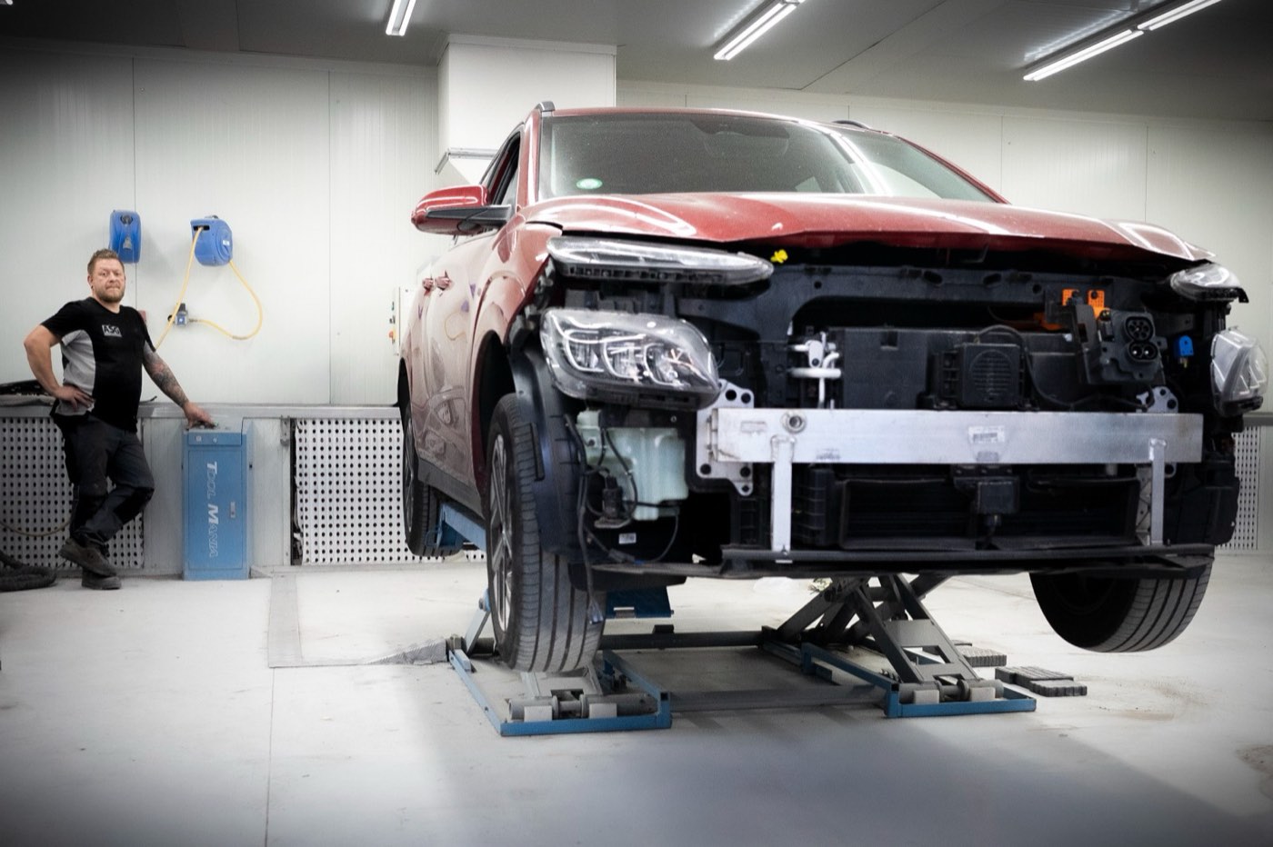 Een rode auto met gedemonteerde voorzijde op een hefbank in de werkplaats, met een monteur ernaast